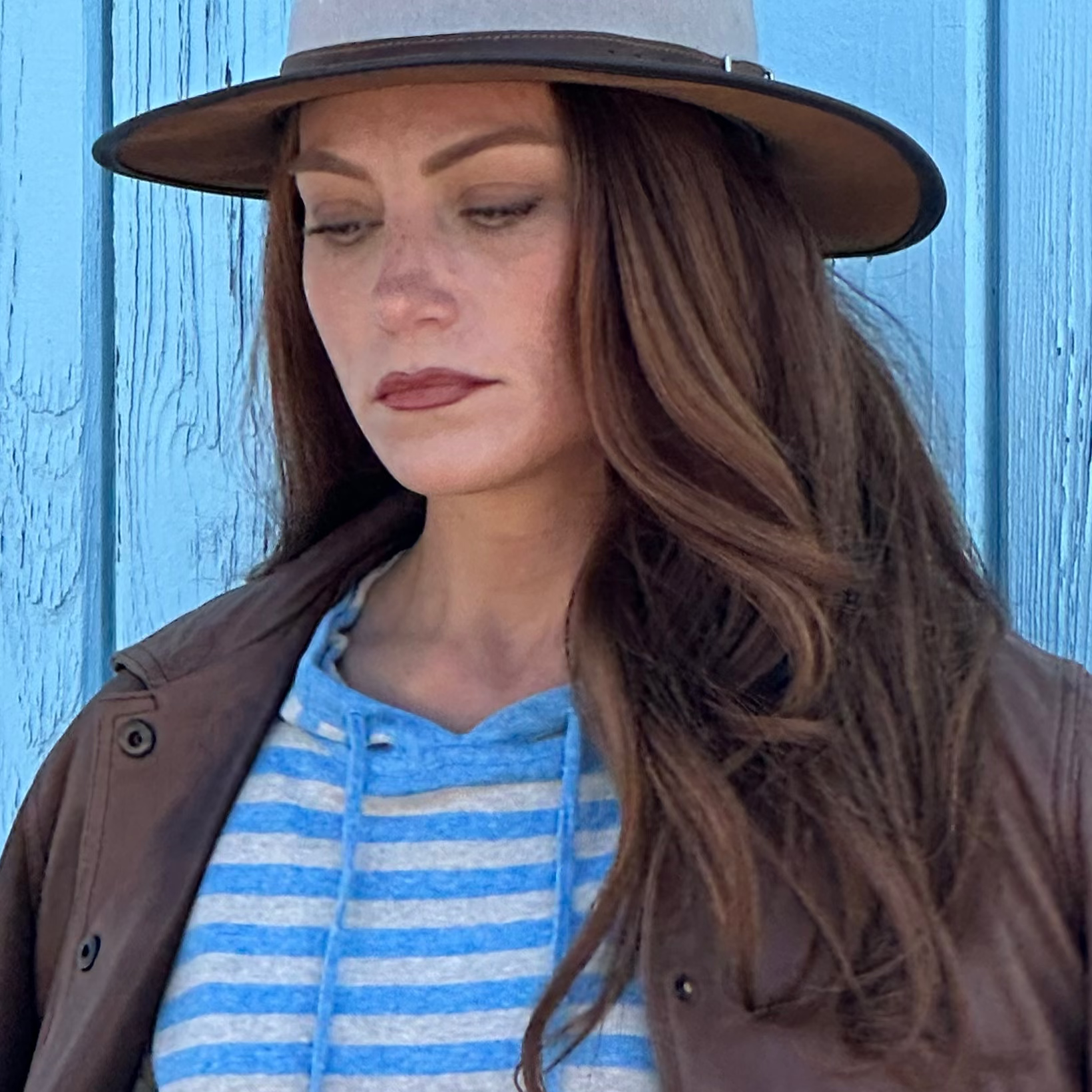 Woman wearing a brown jacket, blue striped hoodie, and light blue jeans against a blue wooden background.