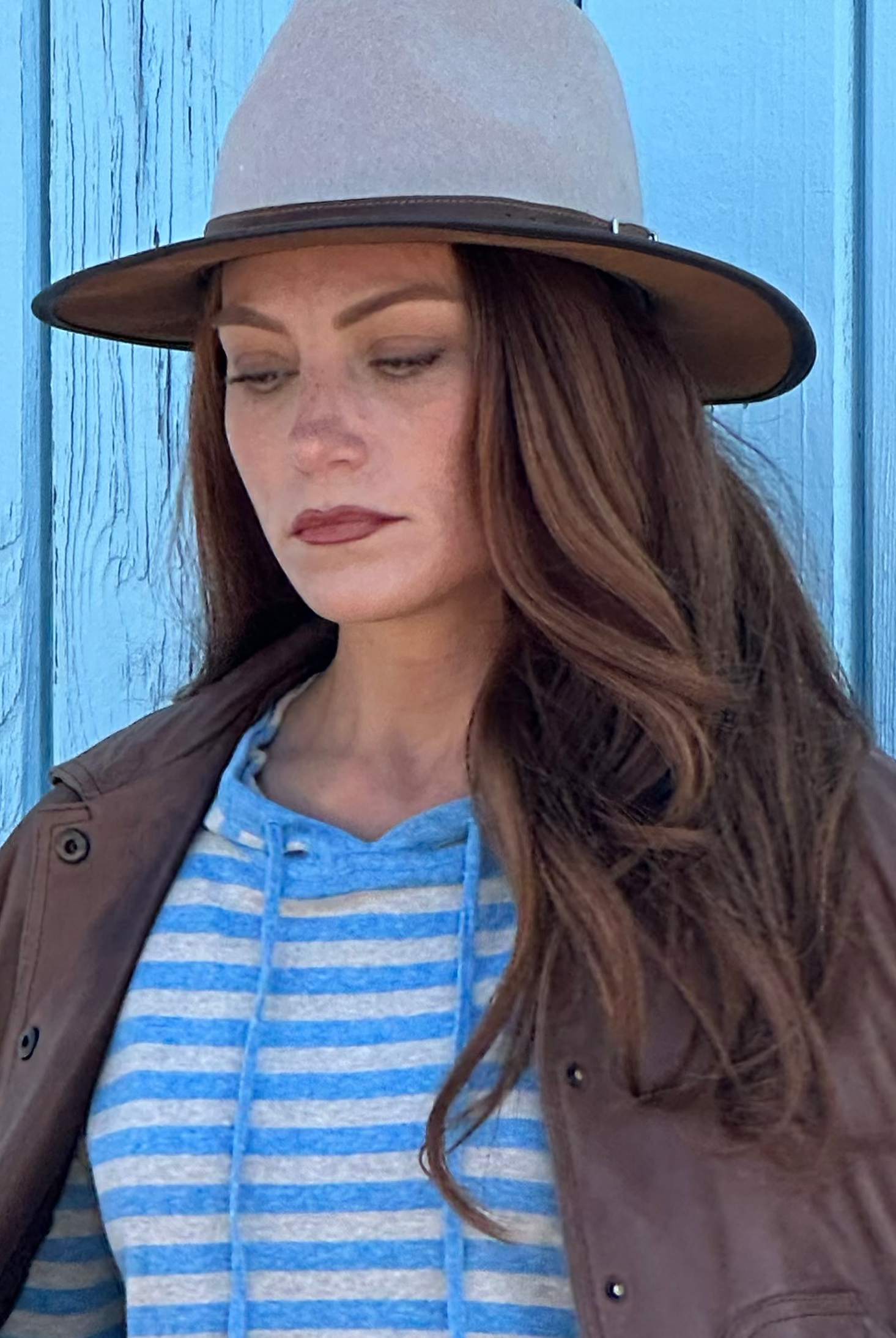 Woman wearing a brown jacket, blue striped hoodie, and light blue jeans against a blue wooden background.