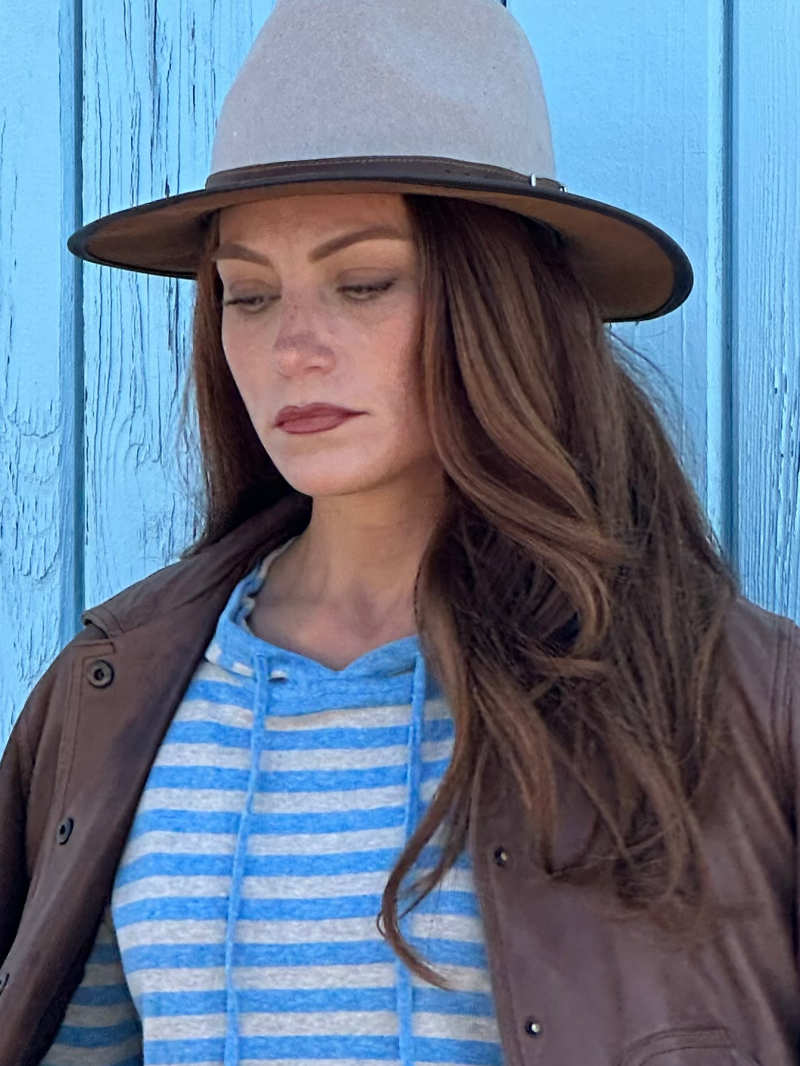Woman wearing a brown jacket, blue striped hoodie, and light blue jeans against a blue wooden background.